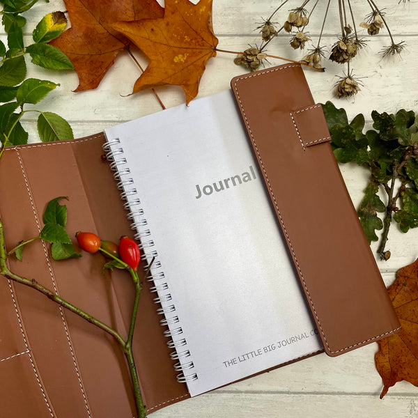 A brown faux leather refillable journal with a magnetic clasp, labelled 'JOURNAL' on the front cover of the refillable insert, it is displayed on a wooden surface surrounded by autumn leaves and flowers.