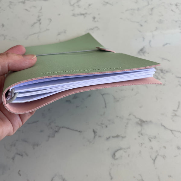 A hand holding a slim, two-tone planner or notebook cover in sage green and blush pink, filled with white pages, set against a light gray marble background.