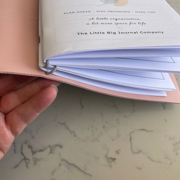 A hand holding an open pink ring-bound journal, showing white pages with printed text and a marble surface underneath.