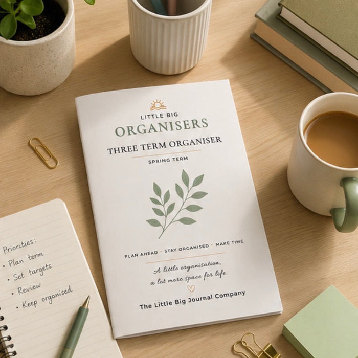 A white spring term planner from The Little Big Journal Company lies open on a light wood desk, surrounded by a coffee cup, notebooks, a small plant, and stationery accessories.