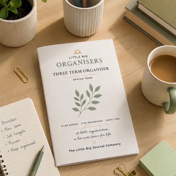 A white spring term planner from The Little Big Journal Company lies open on a light wood desk, surrounded by a coffee cup, notebooks, a small plant, and stationery accessories.