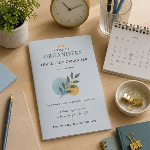 A three-term summer term planner from Little Big Organisers is displayed on a wooden desk surrounded by stationery items including a plant, clock, calendar, pens, and paperclips.