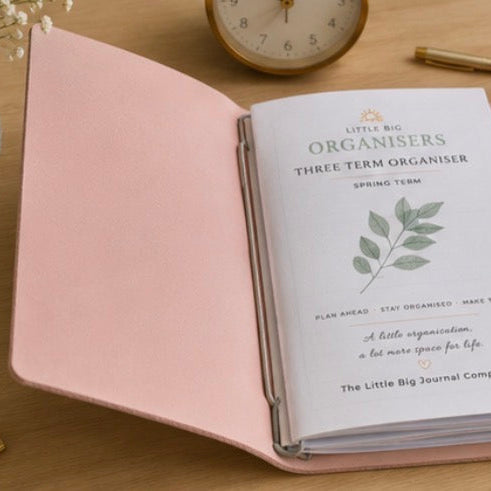 A pink leather-bound three-term spring term planner from The Little Big Journal Company, shown open on a wooden desk with a gold pen, a ceramic mug, a clock, and a binder clip.