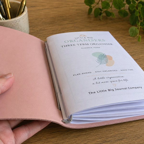 A hand holds an open pink leather planner on a wooden desk. The inside cover shows printed text reading 'Little Big Organisers Three Term Organiser' and includes a small pastel leaf illustration.