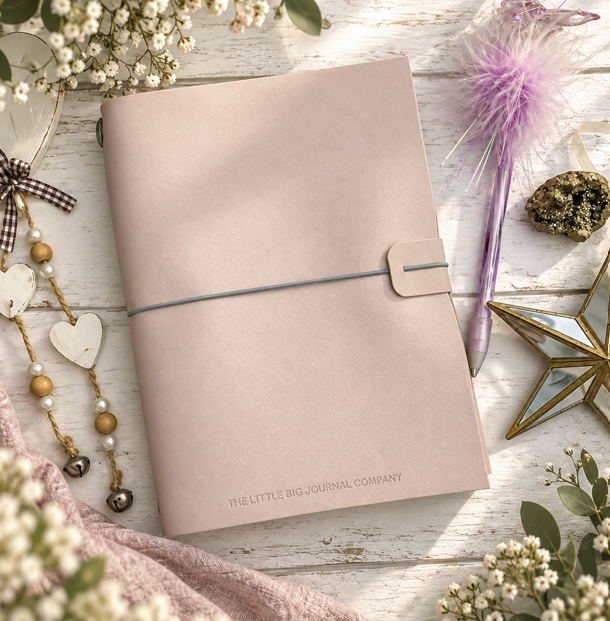 A blush pink journal with a wrap-around strap closure, styled on a white wooden surface with baby's breath flowers, a purple feather pen, and decorative accessories. The journal features the text 'THE LITTLE BIG JOURNAL COMPANY' near the bottom.