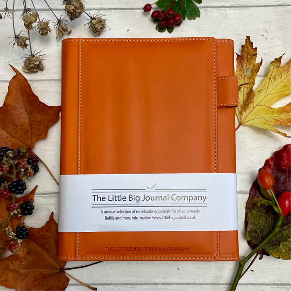 A burnt orange faux leather refillable journal with a magnetic clasp, displayed on a wooden surface surrounded by autumn leaves and berries.